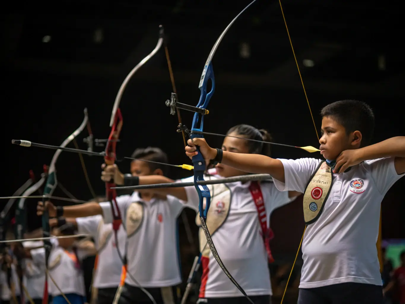 A photograph capturing a beginner archery class in session, with participants learning the basics of stance and aiming under the guidance of an experienced instructor. The setting is the club's indoor training facility.